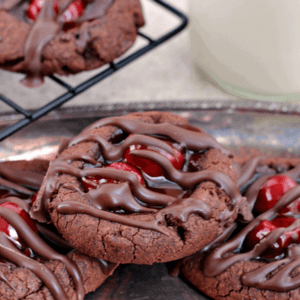 Three chocolate cherry thumbprint cookies on a metal serving tray next to more cookies on a cooing rack and a glass of milk