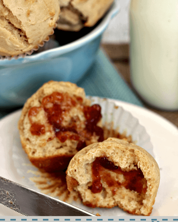 An easy peanut butter muffin cut in half sitting on a cupcake liner in front of a blue bowl of muffins and a glass of milk