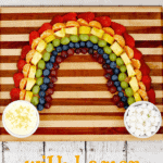 Pot o;gold fruit rainbow on a wood cutting board with a bowl of lemon cheesecake dip at one end of the rainbow and a bowl of mini marshmallows at the other end