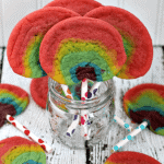 Four rainbow sugar cookie pops in a glass jar surrounded by five rainbow sugar cookie pops on the table