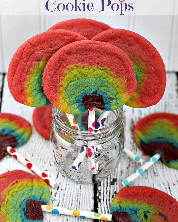 Four rainbow sugar cookie pops in a glass jar surrounded by five rainbow sugar cookie pops on the table