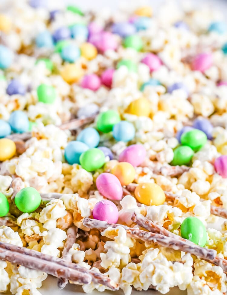 Snack mix spread out on wax paper with popcorn pretzels m&ms and peanuts with candy melts coating