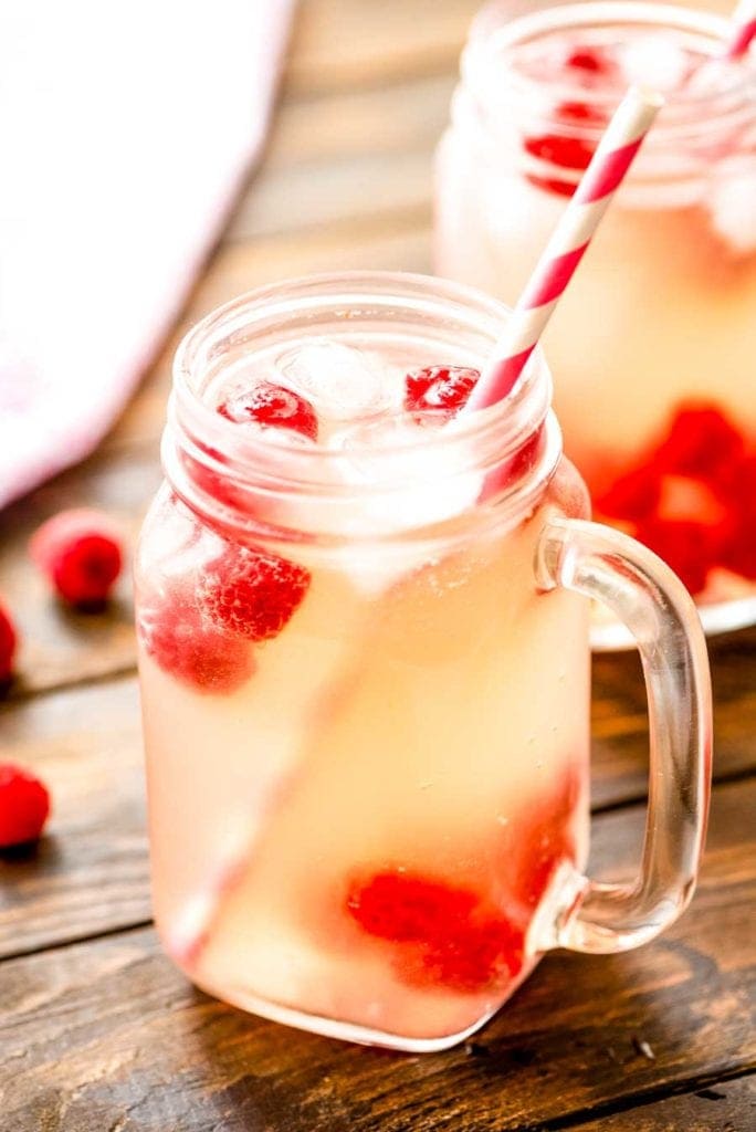 Moscato Lemonade in a mason jar with white and pink striped paper straw on a brown background with raspberries and another mug in the background.