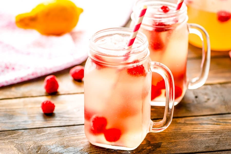 Two mason jars with Pink Moscato Lemonade in them, garnished with white and pink paper straws and raspberries. Sitting on wood background with raspberries and lemons in background.