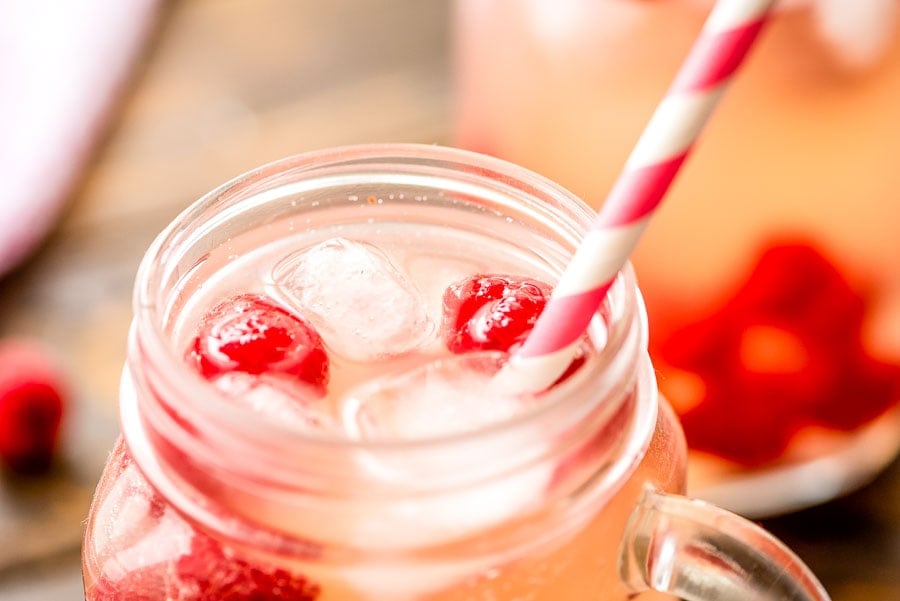 Close up image of mason jar with pink moscato lemonade in it showing only the top. Garnished with raspberries and pink and white striped paper straw.