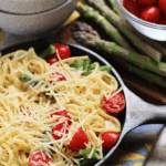 Small cast iron skillet of shrimp and asparagus pasta topped with shredded parmesan on a counter with asparagus and a bowl of cherry tomatoes