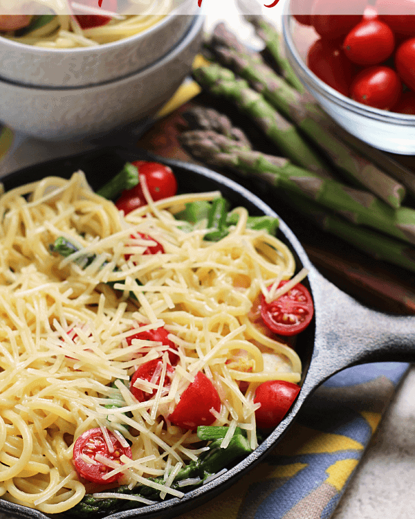 Small cast iron skillet of shrimp and asparagus pasta topped with shredded parmesan on a counter with asparagus and a bowl of cherry tomatoes