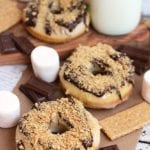 Three s'mores donuts on a table with marshmallows, chocolate bars, graham crackers and a glass of milk