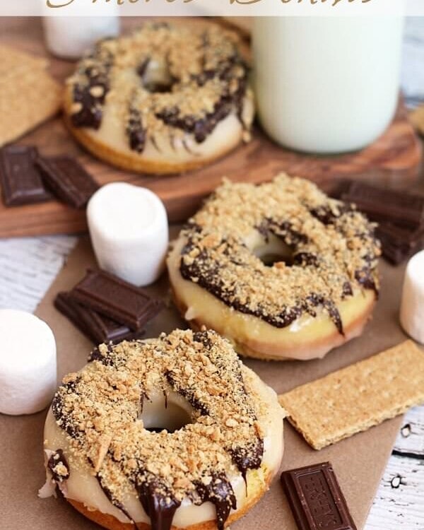 Three s'mores donuts on a table with marshmallows, chocolate bars, graham crackers and a glass of milk