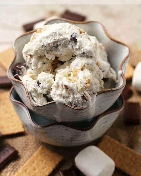 Two brown bowls stacked with the top full of s'mores ice cream and surrounded by graham crackers, chocolate chunks, and marshmallows