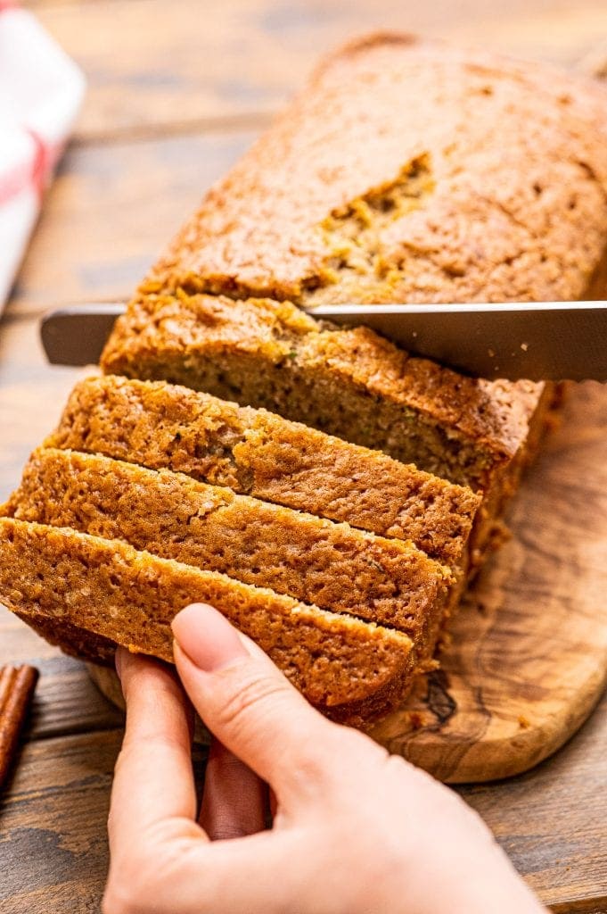 Hand holding a loaf of Banana Zucchini Bread while a knife slices it.