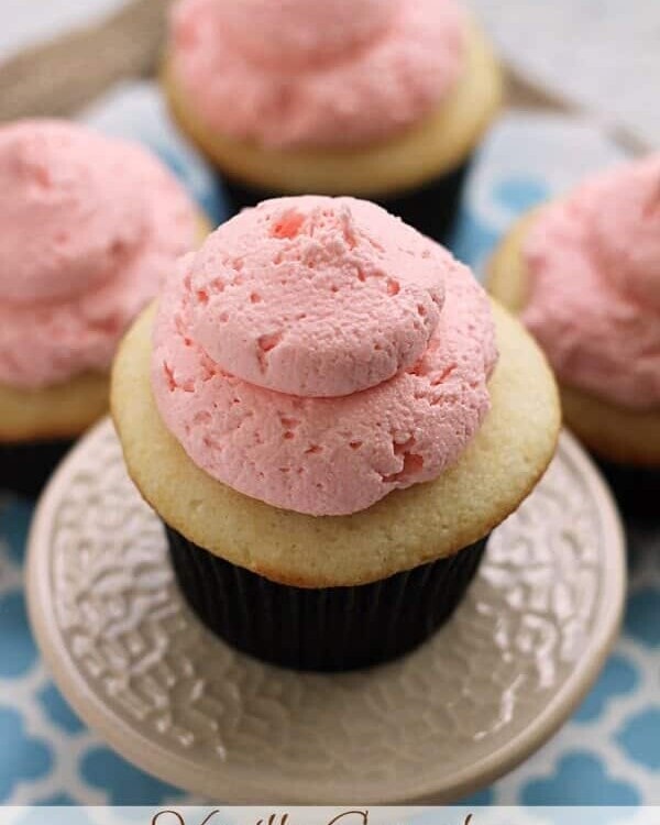 A vanilla cupcake with pink buttercream frosting on a small white cake stand sitting on a blue napkin in front of three more cupcakes