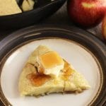 A slice of apple skillet pancake with a pad of butter and syrup on a plate next to a cast iron pan and apples