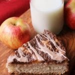 A slice of Cinnamon apple cream cheese coffee cake on a wood board with two apples and a glass of milk