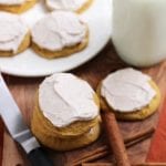 Cinnamon frosted pumpkin cookies, cinnamon sticks, a frosting knife, a glass of milk, and a white plate of cookies on a wood board