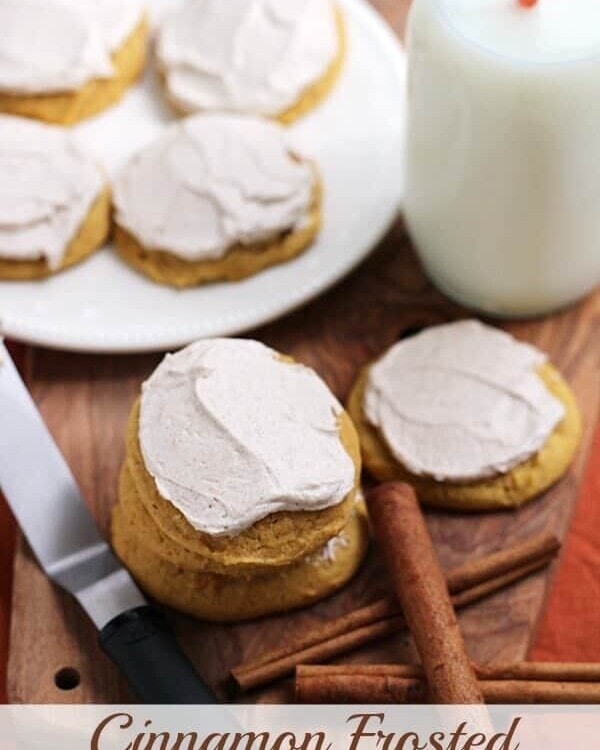 Cinnamon frosted pumpkin cookies, cinnamon sticks, a frosting knife, a glass of milk, and a white plate of cookies on a wood board