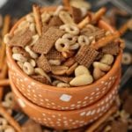 Two orange bowls stacked with the top full of crock pot chex mix and surrounded by chex mix on a napkin