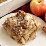 Piece of overnight caramel apple french toast on a white plate next to apples, cinnamon sticks, and a white casserole dish