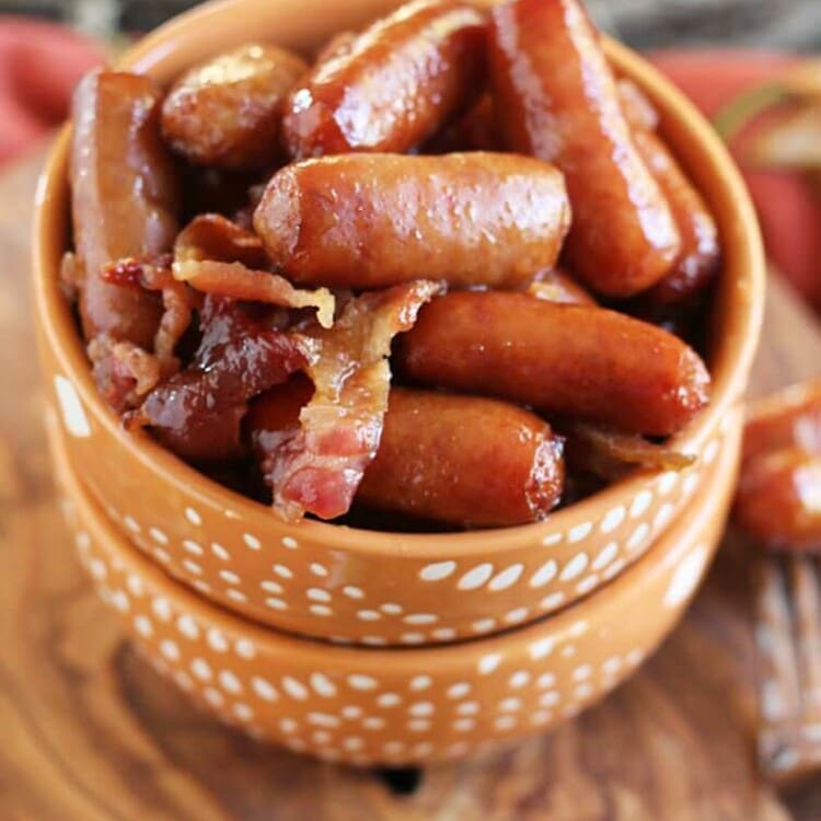 Two orange bowls stacked on a wood board with the top full of crock pot sweet bacon smokies