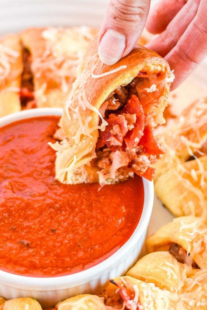 Hand dipping a piece of pizza ring into pizza sauce in bowl