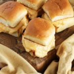 Four cheesy crab sliders on a wood board sitting on a yellow kitchen towel