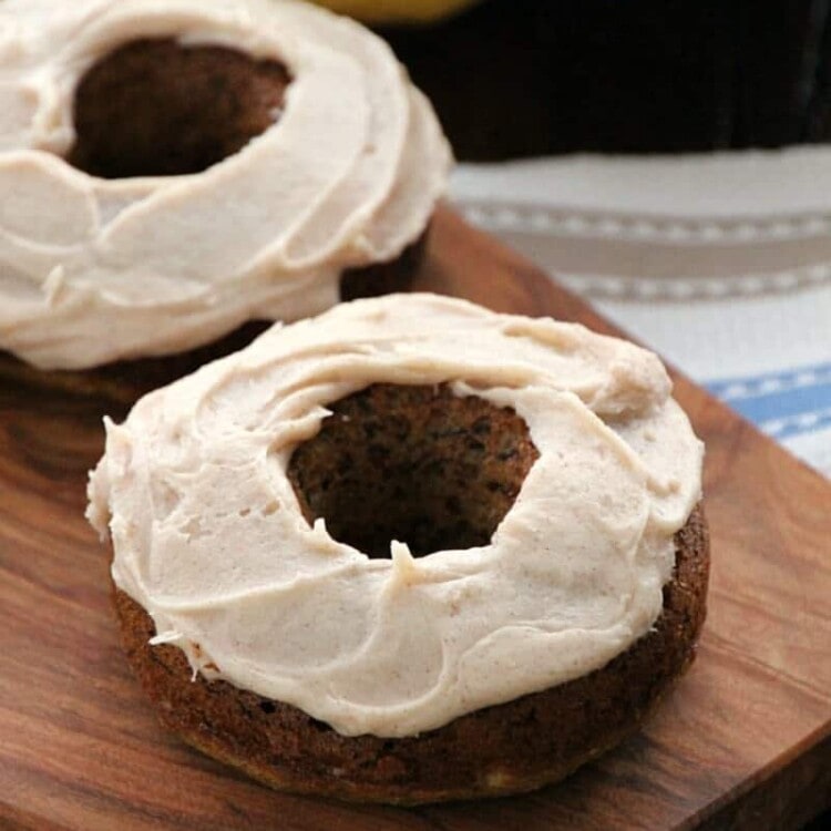 Two cinnamon cream cheese frosted banana donuts on a wood board in front of bananas