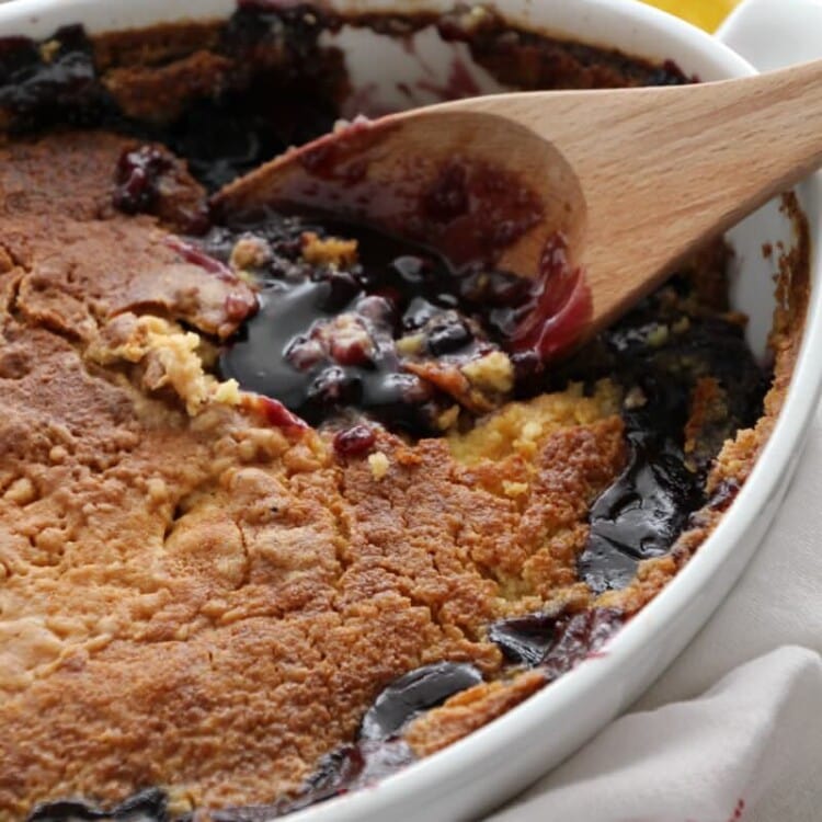 A white oval baking dish full of lemon blueberry dump cake with a wooden spoon next to two lemons