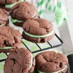 Mint chocolate sandwich cookies on a cooling rack over a mint green napkin next to a jar of green straws and a knife with green frosting on it