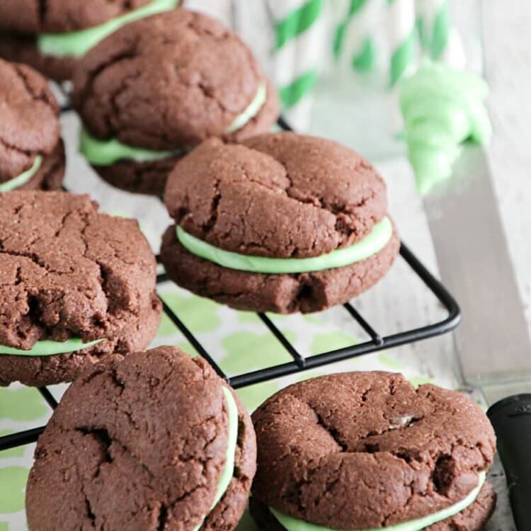 Mint chocolate sandwich cookies on a cooling rack over a mint green napkin next to a jar of green straws and a knife with green frosting on it