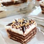 Piece of peanut butter fudge icebox cake on a white square plate sitting on a white table next to a clear glass baking dish of the rest of the cake