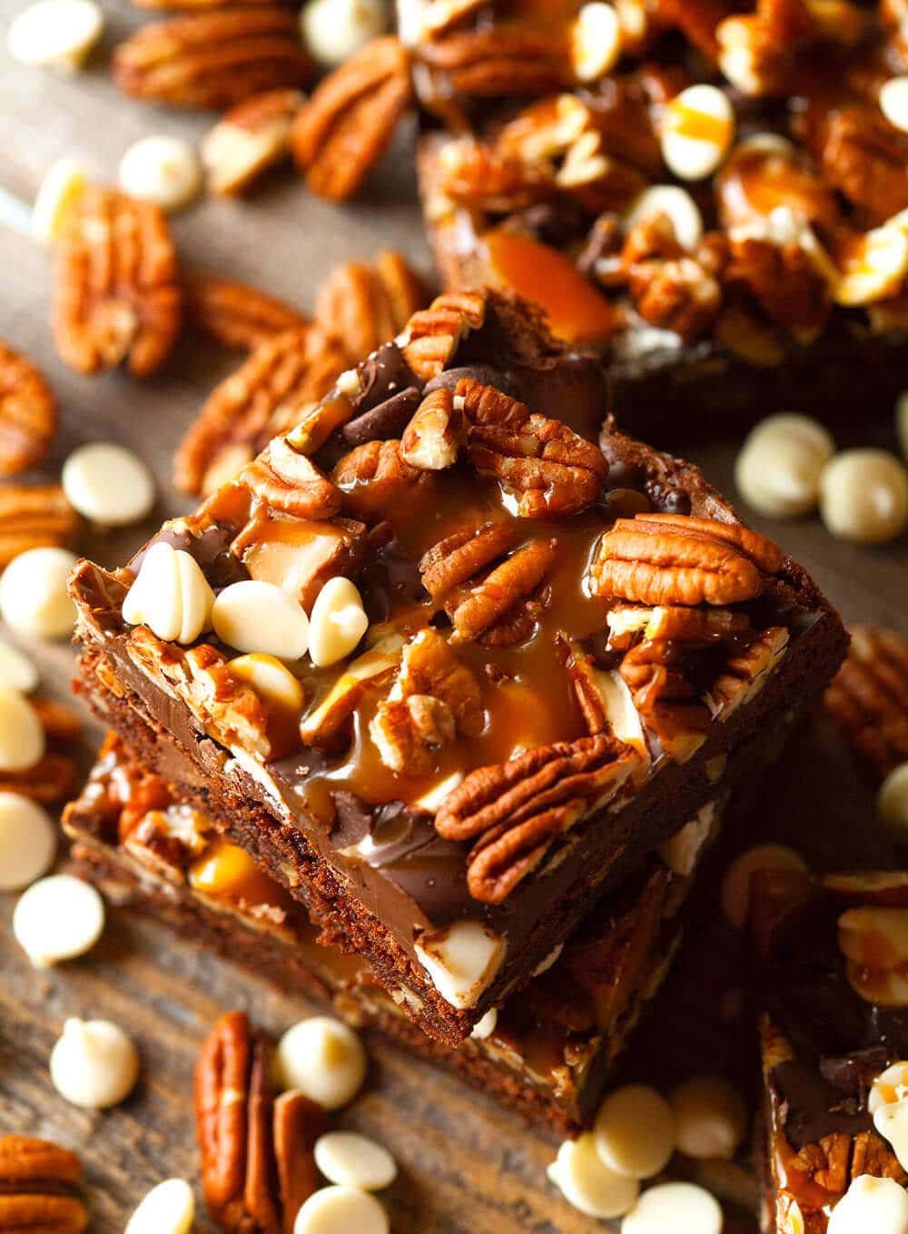 Stack of two turtle brownies with more in the background along with pecans, white chocolate chips on wood board.