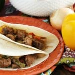 Two crock pot steak fajitas on an orange plate next to a yellow bell pepper, an onion, and a white bowl of steak fajita filling