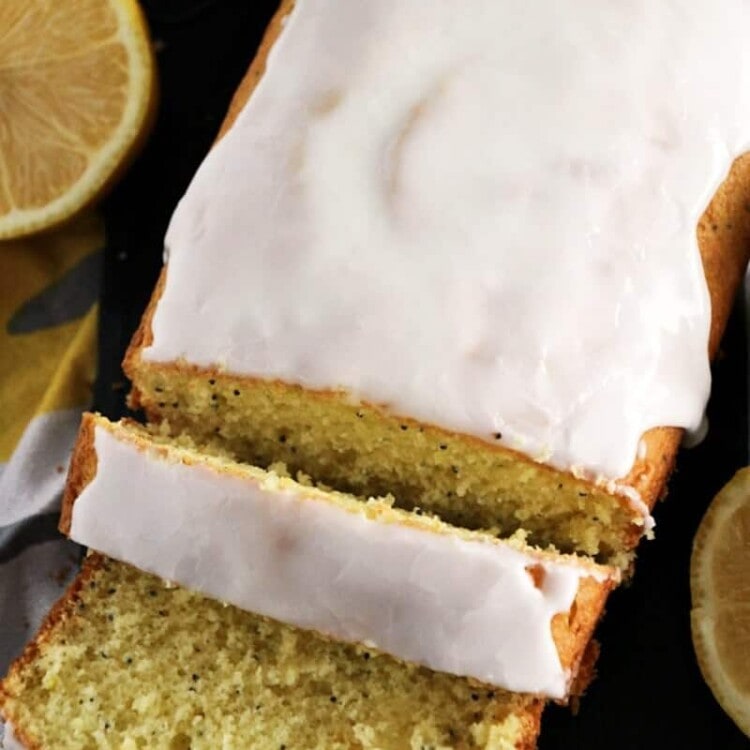 Partially sliced loaf of glazed lemon poppy seed bread next to sliced lemons