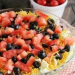 Loaded guacamole dip in a clear glass pie plate next to a white bowl of cherry tomatoes, an orange bowl of corn chips, and an avocado