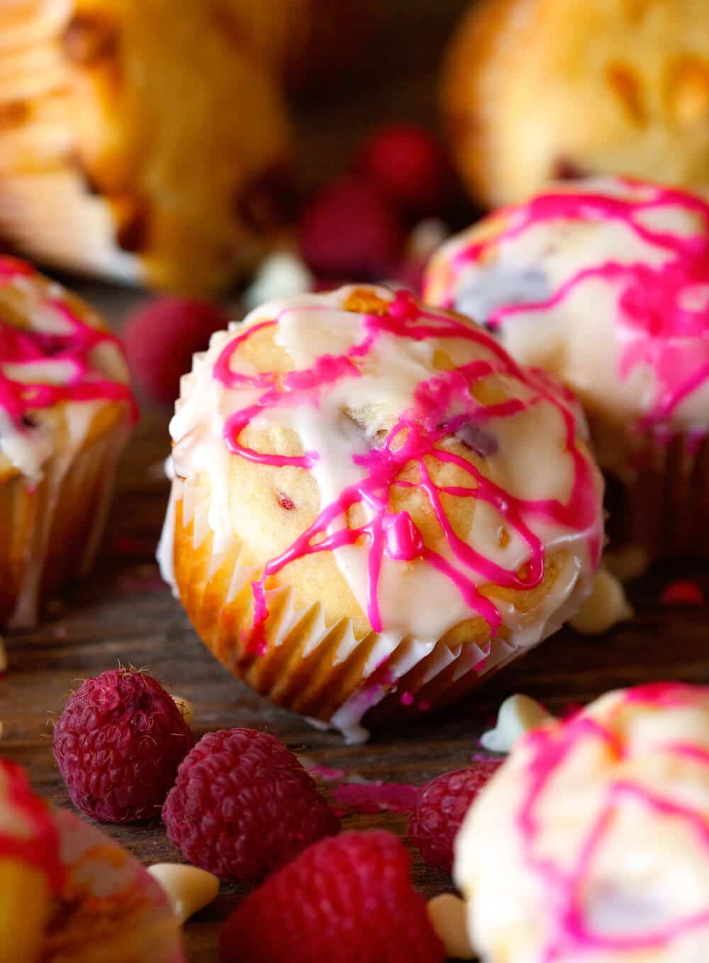 Muffin tilted on its side sitting on wooden background that's topped with pink and white glaze. Fresh raspberries next to it and white chocolate chips.