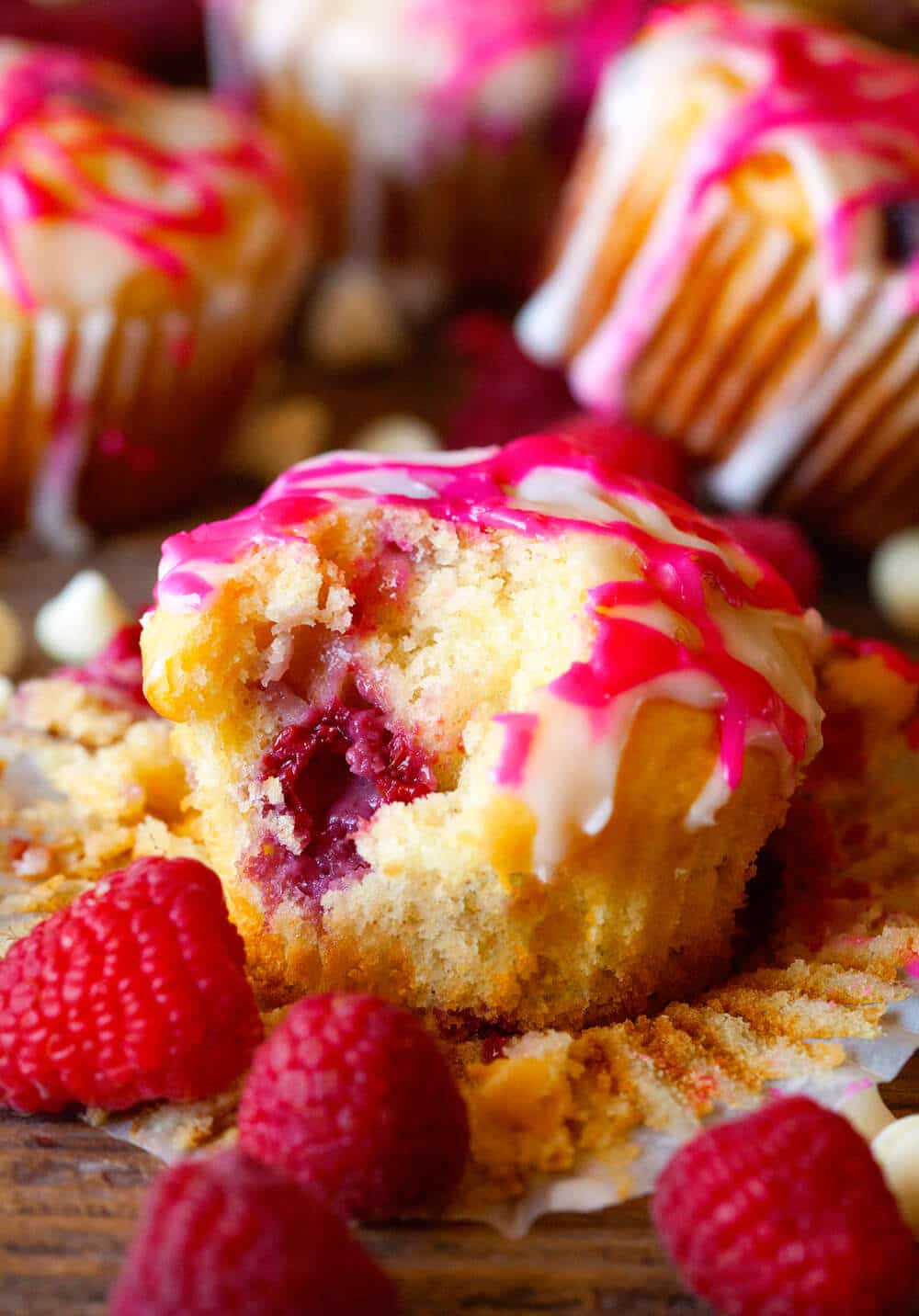 Raspberry Muffin with glaze that is sitting on it's muffin liner after being unwrapped and a bite taken out of it. Fresh raspberries and white chocolate chips next to muffin on wooden background.