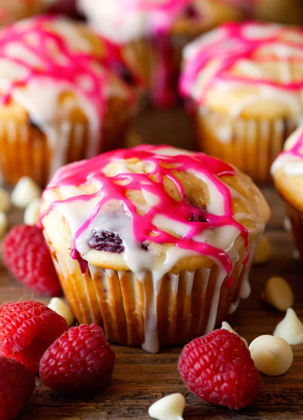 Raspberry Muffin on wooden background topped with white and pink glaze. Fresh raspberries and white chocolate chips laying next to muffins.
