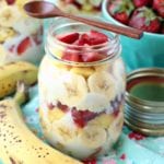 Three mason jars of banana split trifle on a blue table cloth with bananas and a blue bowl of strawberries