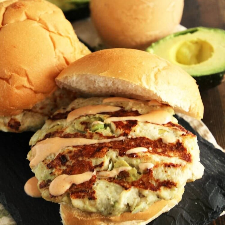 A chicken avocado burger on a bun with chipotle yogurt sauce on a slate tray next to a glass jar of chipotle yogurt sauce and an avocado sliced in half