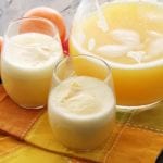 Two glasses and a glass pitcher of mimosa float sitting on an orange cloth napkin