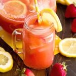 A mason jar mug and a glass pitcher of spiked strawberry lemonade on a wood table with strawberries and lemon slices