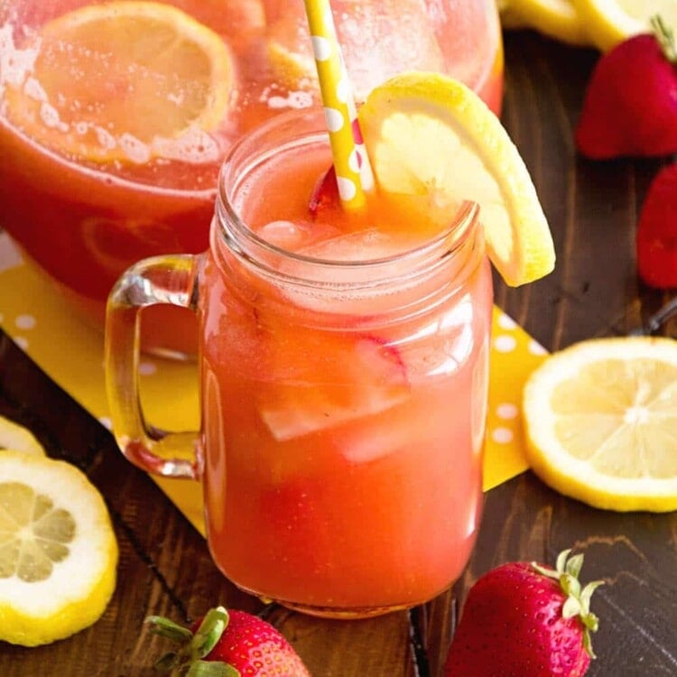 A mason jar mug and a glass pitcher of spiked strawberry lemonade on a wood table with strawberries and lemon slices