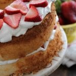 A strawberry lemon fluff angel food cake on a white cake stand in front of a yellow bowl of strawberries