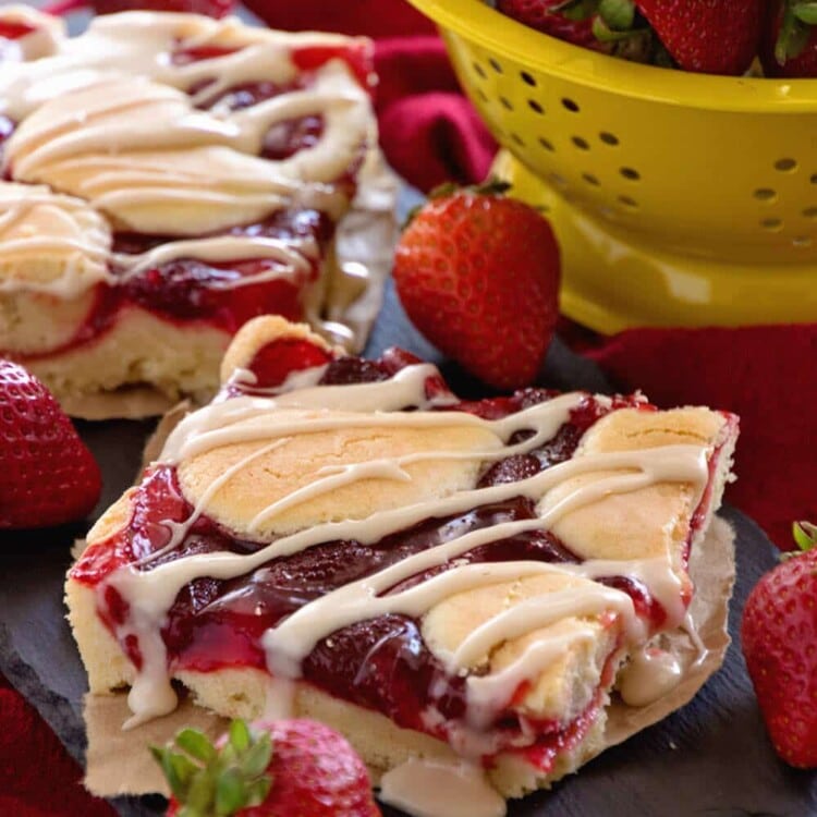 Two strawberry pie bars on a slate tray sitting on a red kitchen towel with a yellow strainer of strawberries
