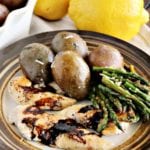 A plate of balsamic chicken, asparagus, and potatoes on a white kitchen towel next to two lemons and a white bowl of potatoes
