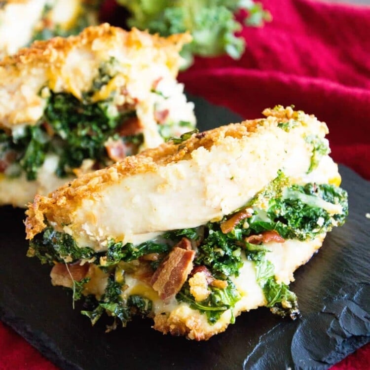 Three cheesy bacon kale stuffed chicken breasts on a slate tray sitting on a red kitchen towel