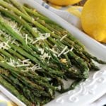 Lemon roasted asparagus topped with shredded parmesan on a white plate next to two lemons