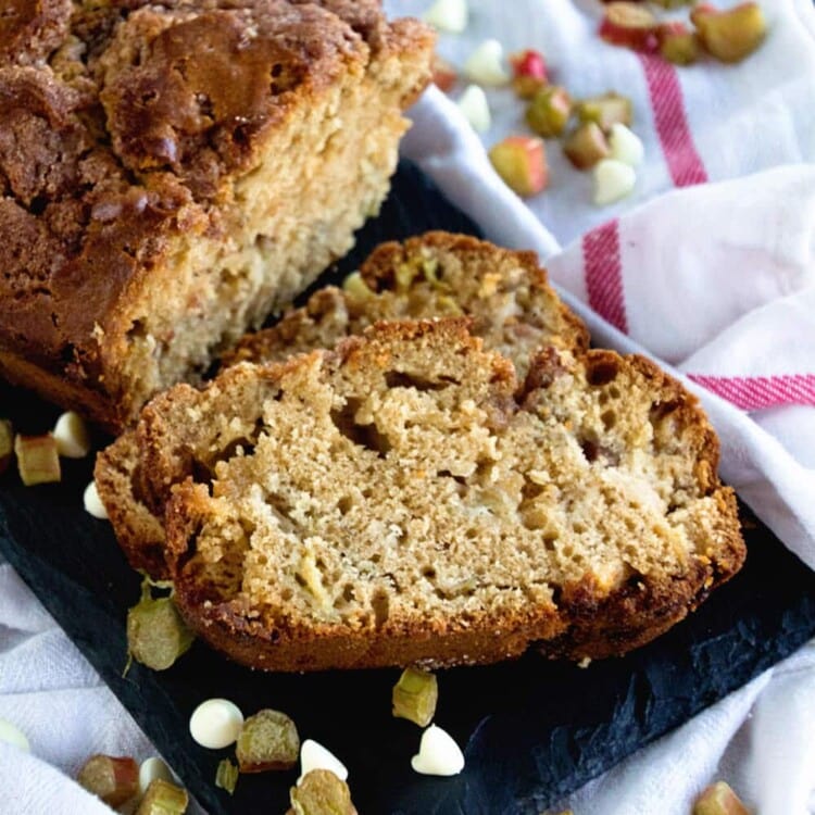 Two slices of buttermilk rhubarb bread cut off of the loaf of bread on a slate tray scattered with white chocolate chips and pieces of rhubarb