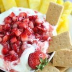 Overhead image of a white bowl of strawberry cheesecake dip with graham crackers and pineapple around it