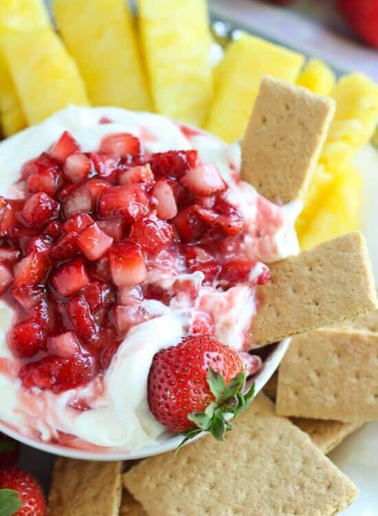 Overhead image of a white bowl of strawberry cheesecake dip with graham crackers and pineapple around it